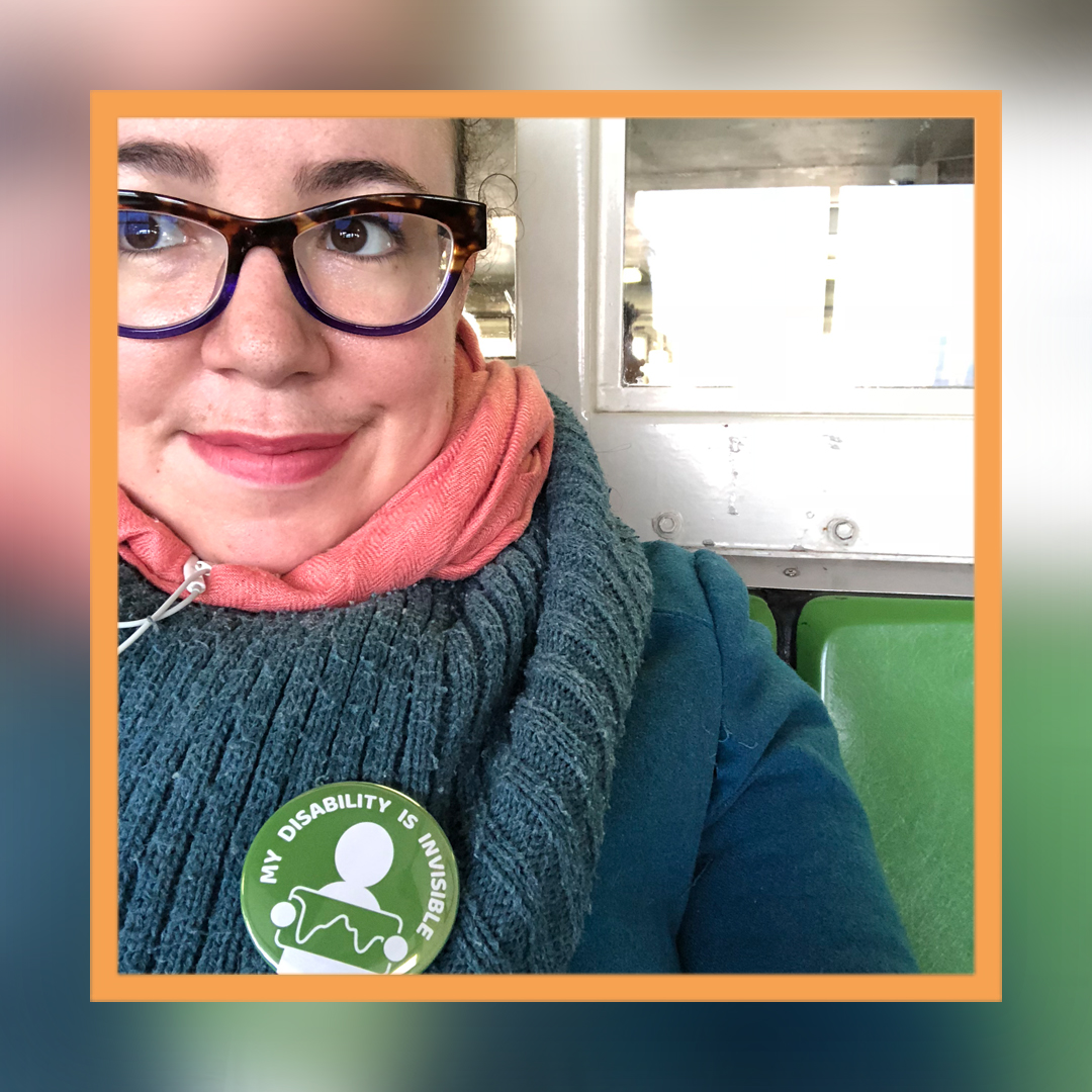 A close-cropped photo of Tema's face and shoulders. She is wearing tortoise shell glasses and looking off camera, and wearing a thick wool scarf with a green button that reads "My disability is invisible."