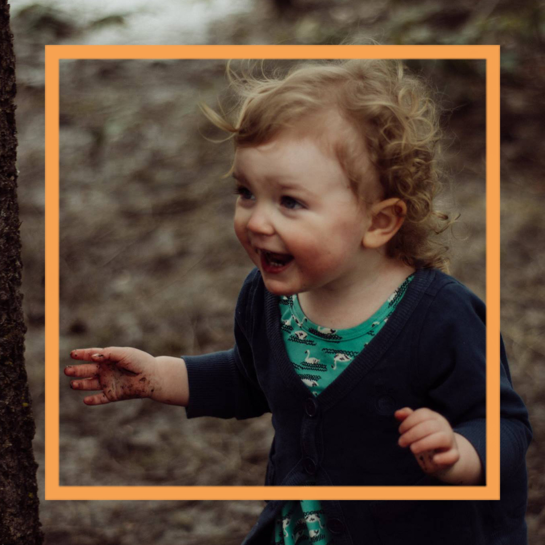 A photo of India, a 2-year-old girl, standing in the woods and smiling off camera.