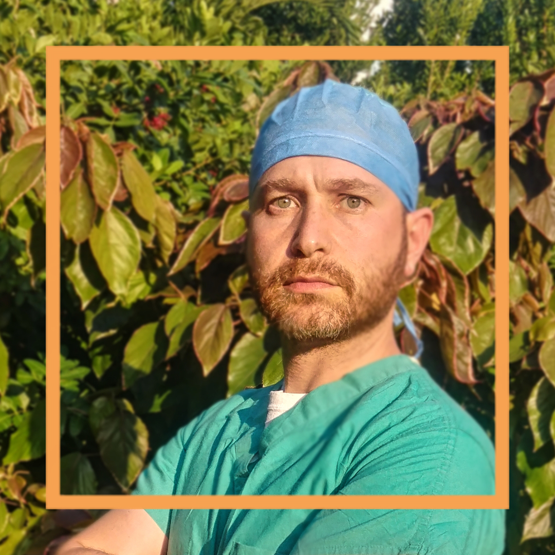 Jacob is standing in front of a shrub, wearing medical scrubs and looking at the camera.