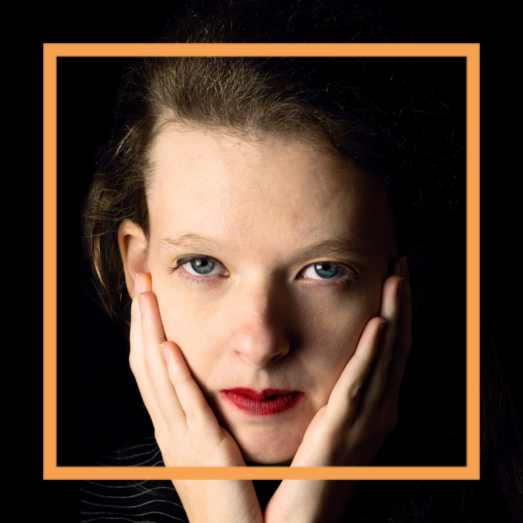 Belle is wearing bright red lipstick and looking at the camera while she rests her chin on her hands. There is an orange box superimposed around her face.