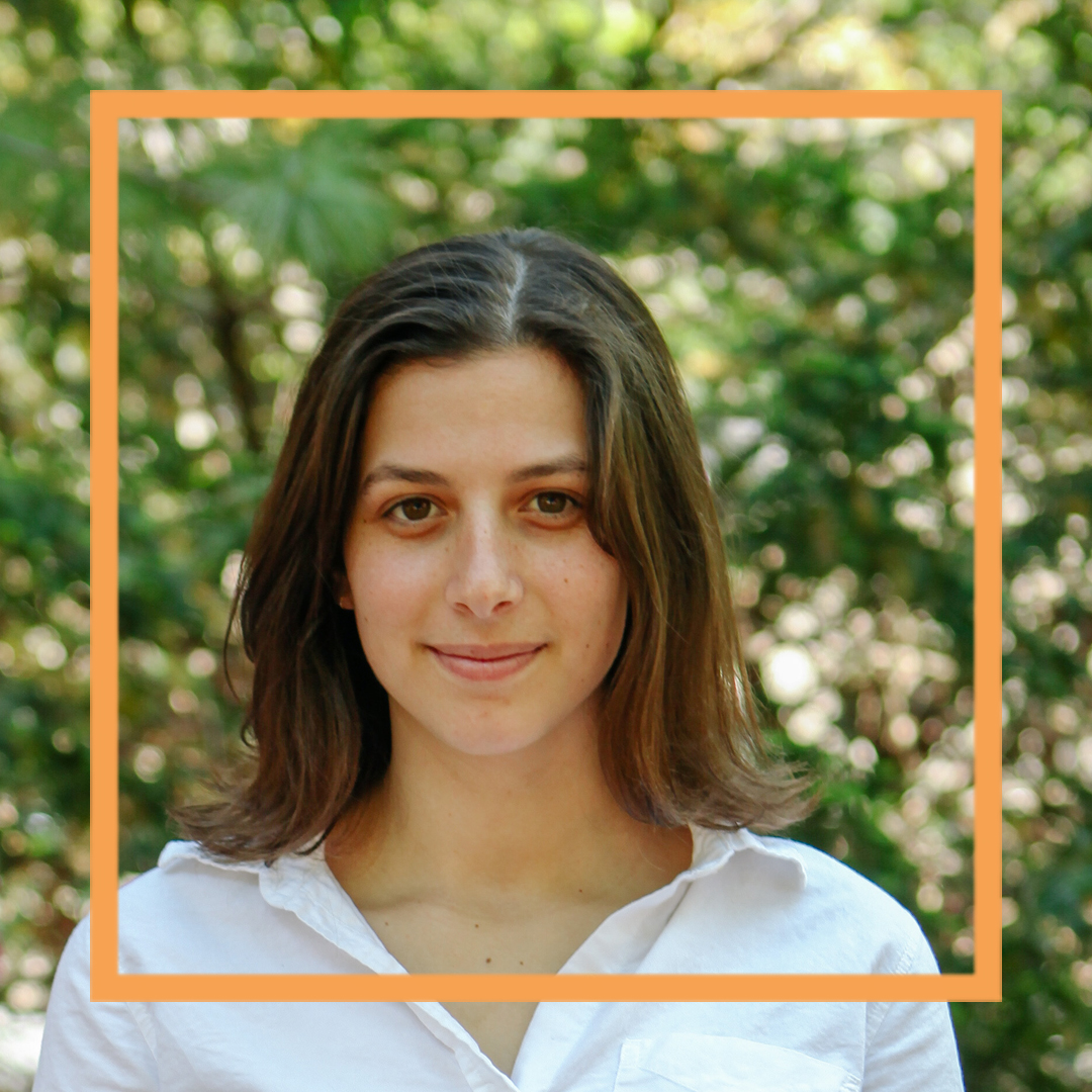 Hannah is wearing a white button-up shirt and standing in front of blurry greenery. She is looking directly at the camera and smiling. There is an orange box superimposed over the photo.