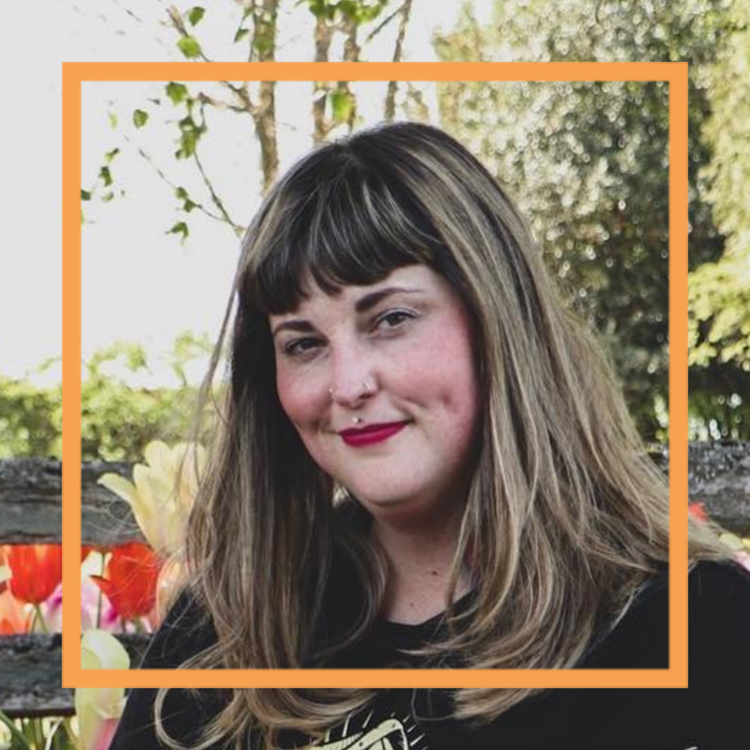 Vanessa, a woman with blunt cut bangs and long highlighted hair, is wearing red lipstick and smiling in front of the camera. There are leaves in the background and there's an orange box superimposed above her face.