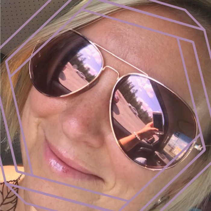 A closeup up of Chrystal smiling at the camera and wearing aviator sunglasses. There is a stylized purple hexagon framing the photo.