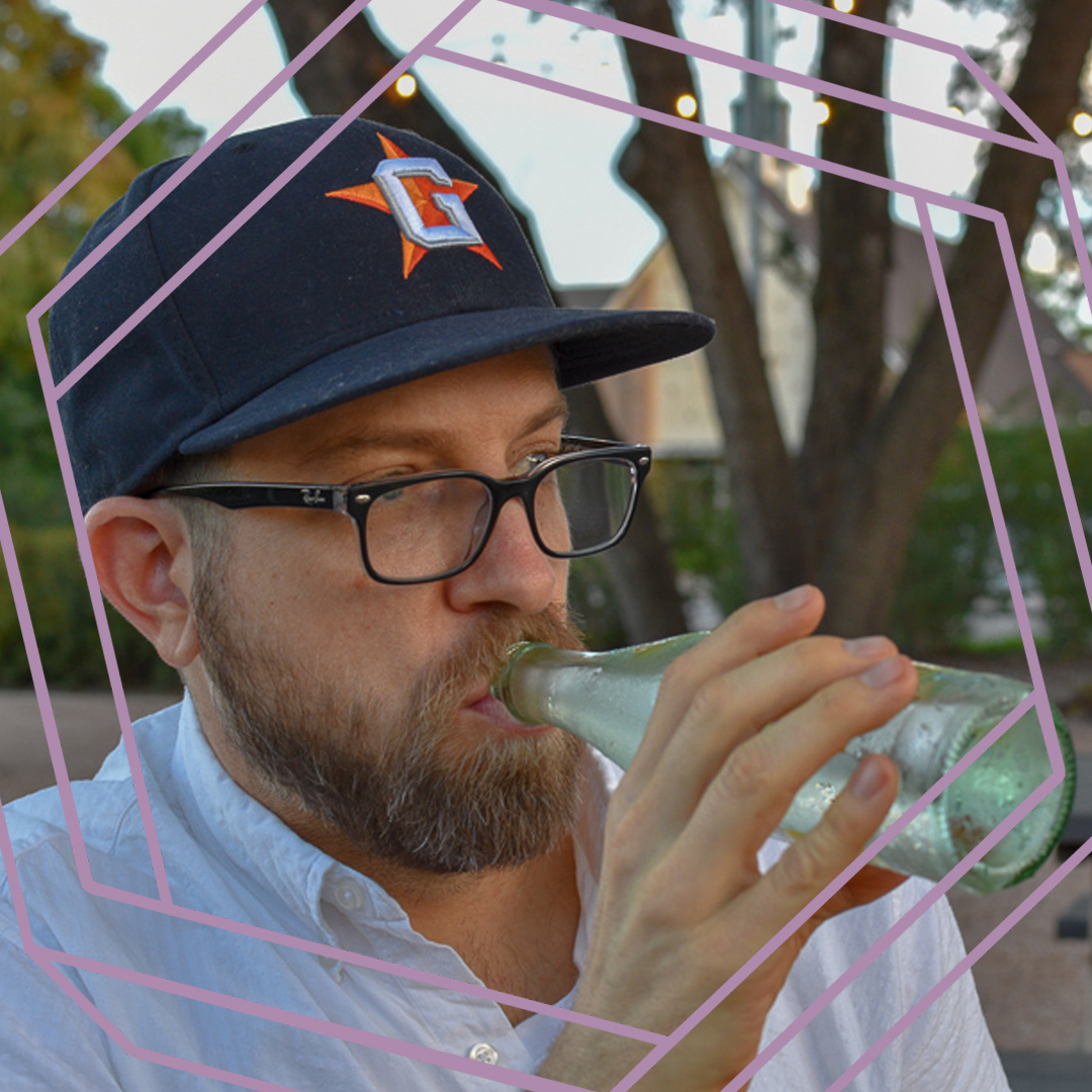 Garreth, a man with a beard, black framed glasses and a navy blue baseball cap, is drinking from a glass bottle and looking off camera. There is a stylized purple octagon superimposed over the photo.