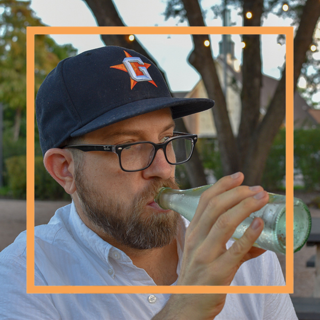 Garreth, a man with a beard, black framed glasses and a navy blue baseball cap, is drinking from a glass bottle and looking off camera. There is an orange square superimposed over the photo.