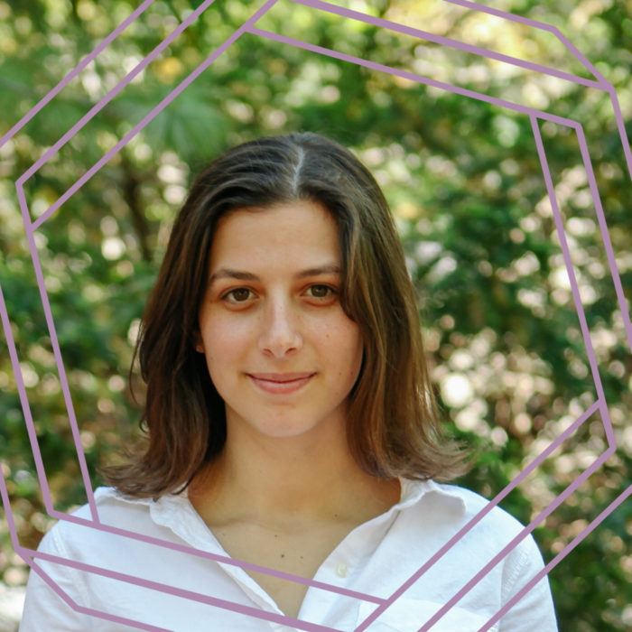 Hannah is wearing a white button-up shirt and standing in front of blurry greenery. She is looking directly at the camera and smiling. There is a stylized purple octagon superimposed over the photo.
