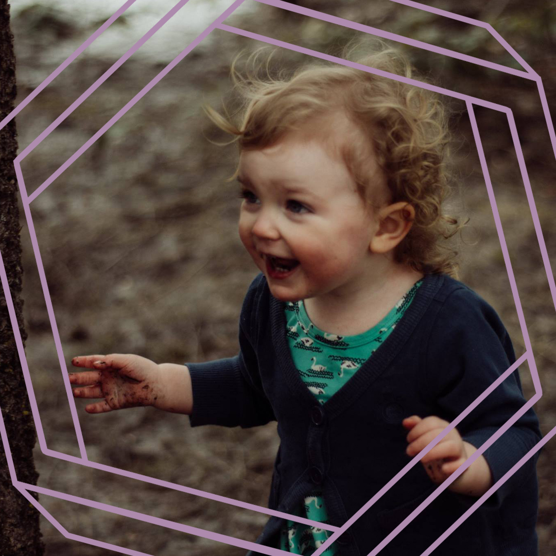 A photo of India, a 2-year-old girl, standing in the woods and smiling off camera. There is a stylized purple hexagon superimposed over the photo.