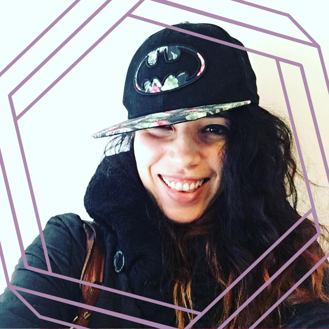 Nicole wears a flatbrimmed black cap with the batman logo and smiles at the camera. There is a stylized purple hexagon framing the photo.