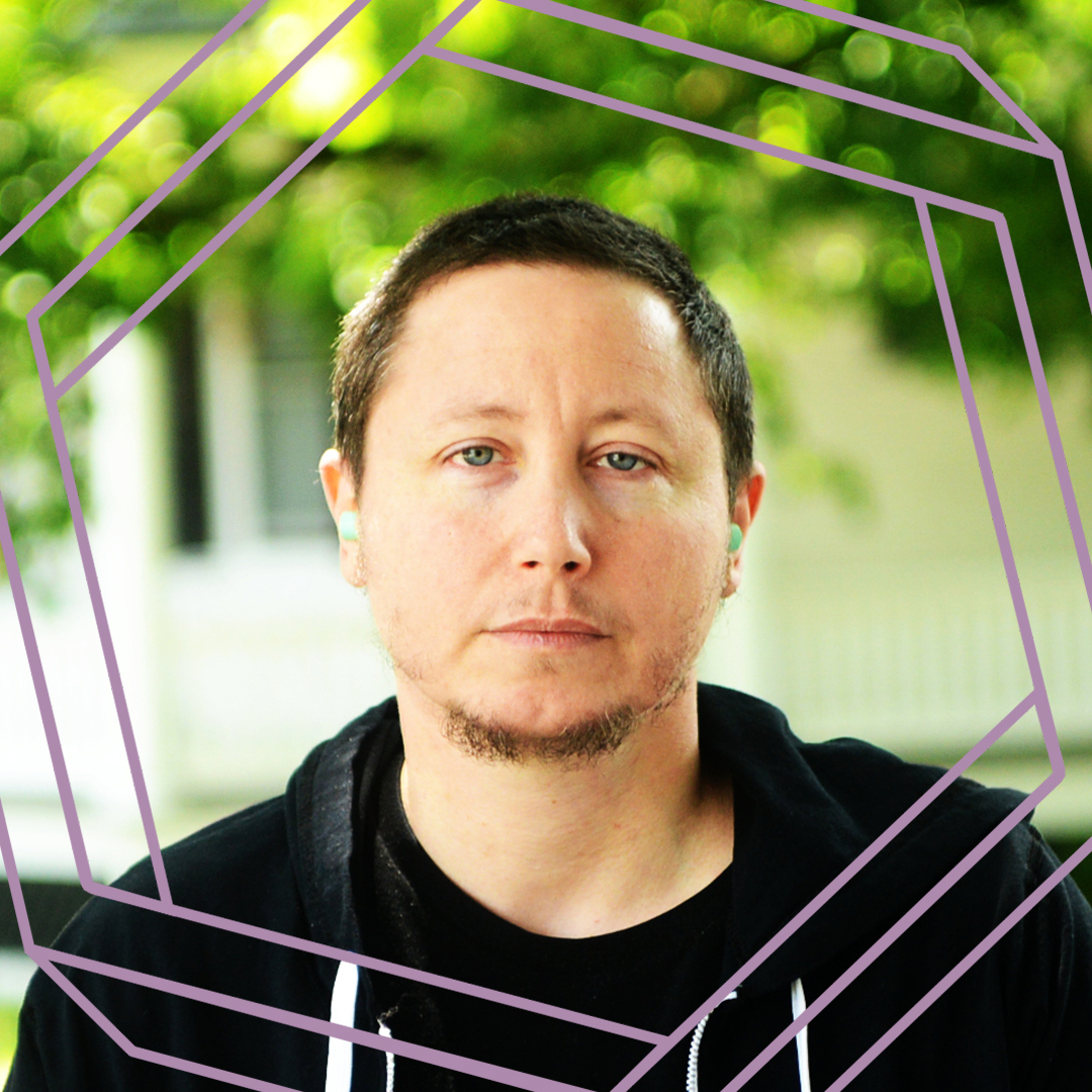 Dov, a white man with short dark hair and a goatee, looks directly at the camera. He is wearing a black hoodie and stands in front of a blurry green background. There is a stylized purple octagon superimposed over the photo.