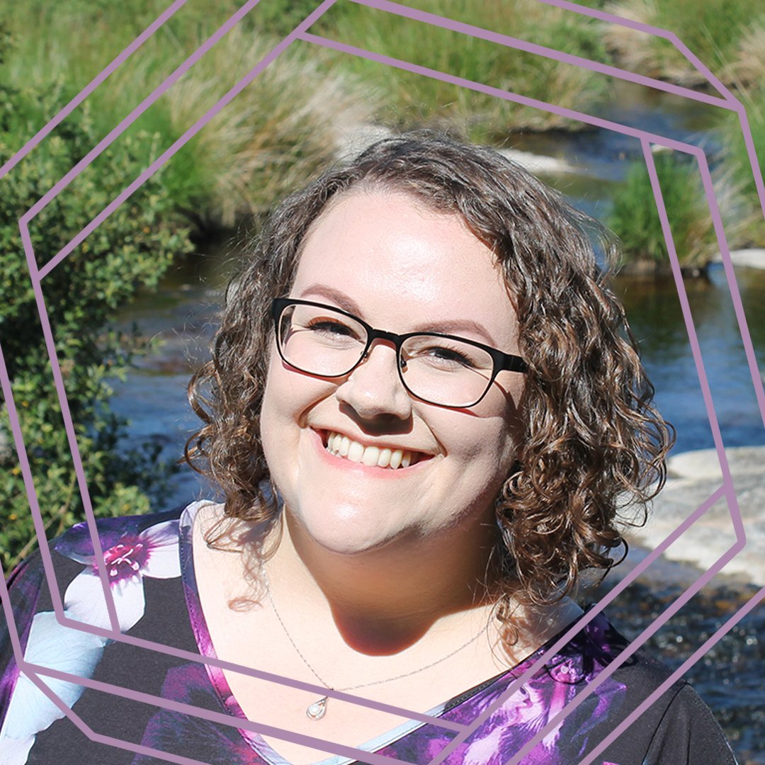 Sarah, a white woman with shoulder-length curly brown hair, smiles at the camera. There is a stream with grassy banks behind her and a stylized purple octagon superimposed over the photo.