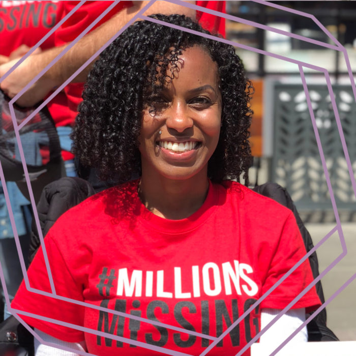 Ashanti, a black woman with curly shoulder-length black hair, smiles at the camera. She is wearing a red shirt that reads "#Millions Missing." There is a purple hexagon superimposed over the photo.