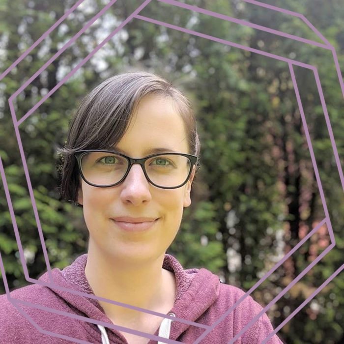 Brianne, a white woman with short brown hair and black plastic framed glasses is smiling at the camera. She is wearing a purple hooded sweatshirt and standing in front of a green hedge background. There is a stylized purple hexagon framing the photo.