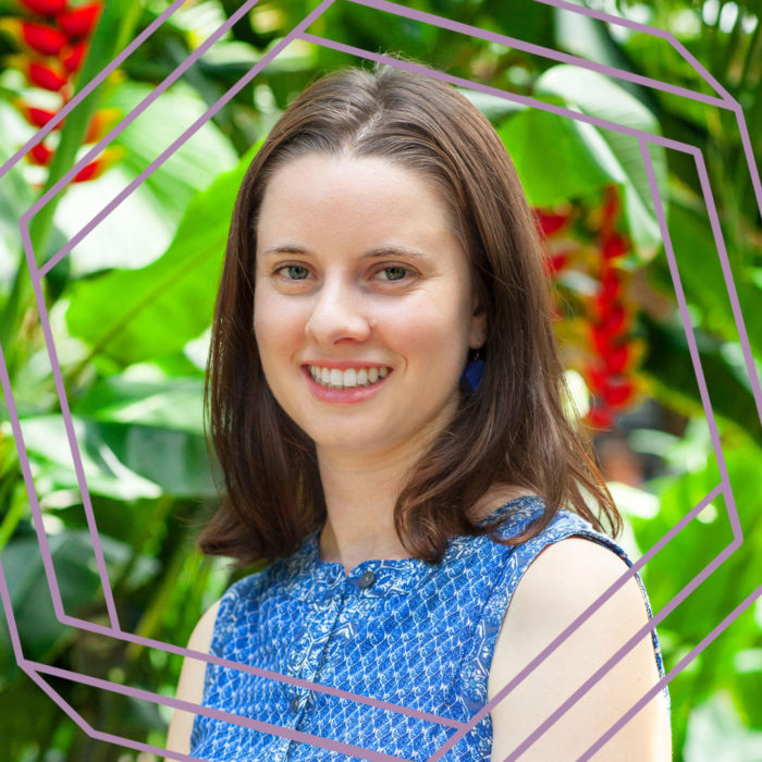 Noa, a white woman in her 20s with shoulder length brown hair, is smiling at the camera. She is wearing a sleeveless blue dress and standing in front of of blurry tropical plants. There is a stylized purple hexagon framing the photo.