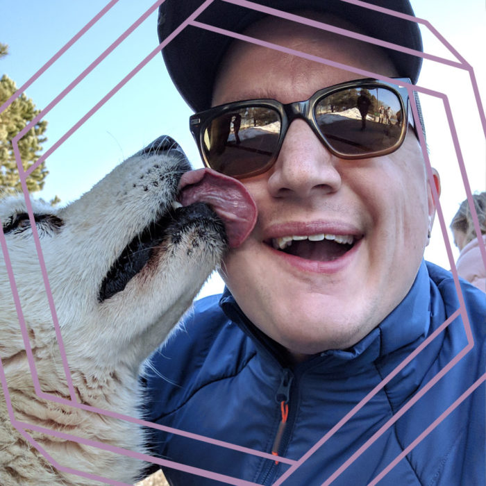 Steve, a white man wearing sunglasses and a ball cap, smiles at the camera while a yellow dog licks his cheek. There is a stylized purple hexagon framing the photo.