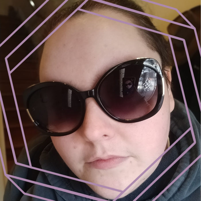 Photo of Emma, a white woman wearing large sunglasses, looking at the camera. There is a stylized hexagon framing the photo.