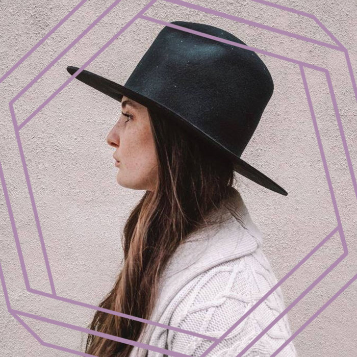 A photo of Taryn in profile.Taryn is a white woman with long brown hair wearing a wide-brimmed black hat and a cable-knit sweater. There is a stylized purple hexagon framing the photo.