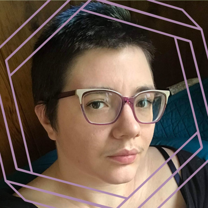 Suzanne, a white person with short brown hair and plastic-framed glasses, looks a the camera. There is a stylized purple hexagon framing the photo.