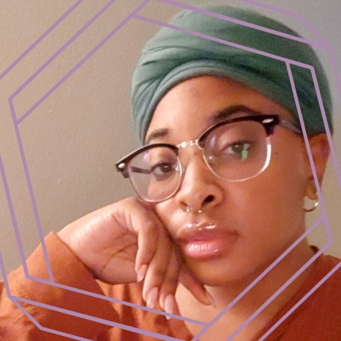 Teona, a Black nonbinary woman in her 20s, looks into the camera with a relaxed expression. She is wearing wire-framed glasses and a silver septum ring. Her head is leaning on her hand, and she wears a rust orange shirt and green headwrap. The photo is framed by a stylized purple hexagon.