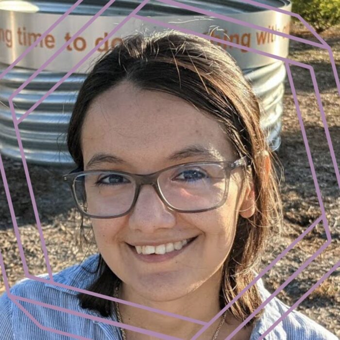 Krystal Vasquez is a light-skinned Latina woman wearing a blue button-up shirt and gray-framed glasses. She is sitting on a bench in a park.The photo is framed by a stylized purple hexagon.