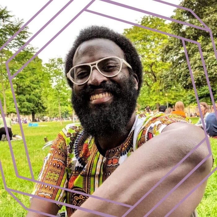 Mugabi, a black luscious full bearded human with white wide-framed glasses and a 4b mini-afro fighting against a receding hairline, smiles in the park with a colourful kintengi print shirt. The photo is framed by a stylized purple hexagon.